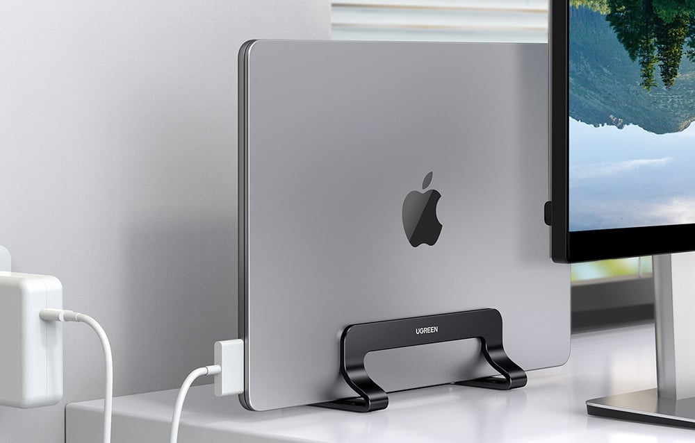 MacBook w czarnej podstawce UGREEN obok monitora i ładowarki na nowoczesnym białym biurku