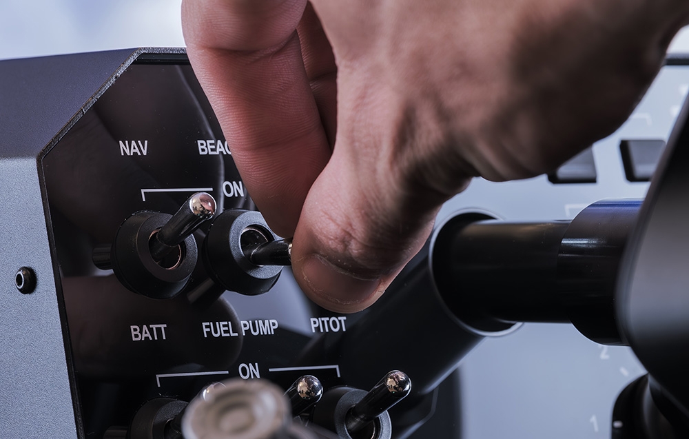 Dłoń osoby obsługującej bazę z napisami NAV, BATT, FULET PUMP, PITOT i ON.