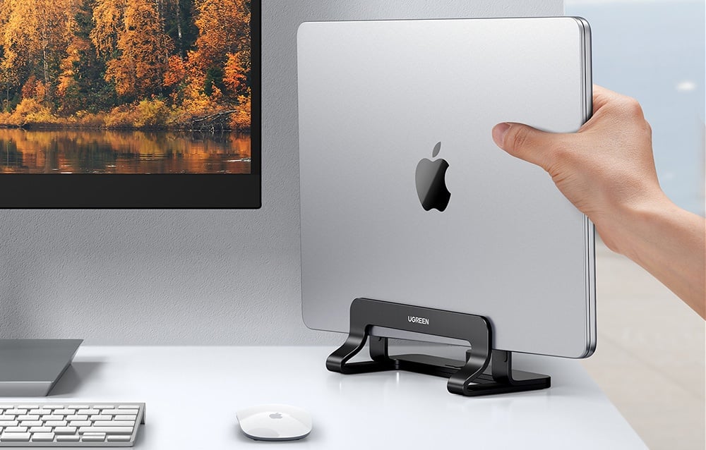 Ręka umieszcza MacBooka w czarnej podstawce UGREEN przy biurku z monitorem i klawiaturą